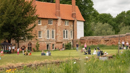 The stone built Charterhouse building forms the backdrop to a scene of families enjoying being outside on a lawn.